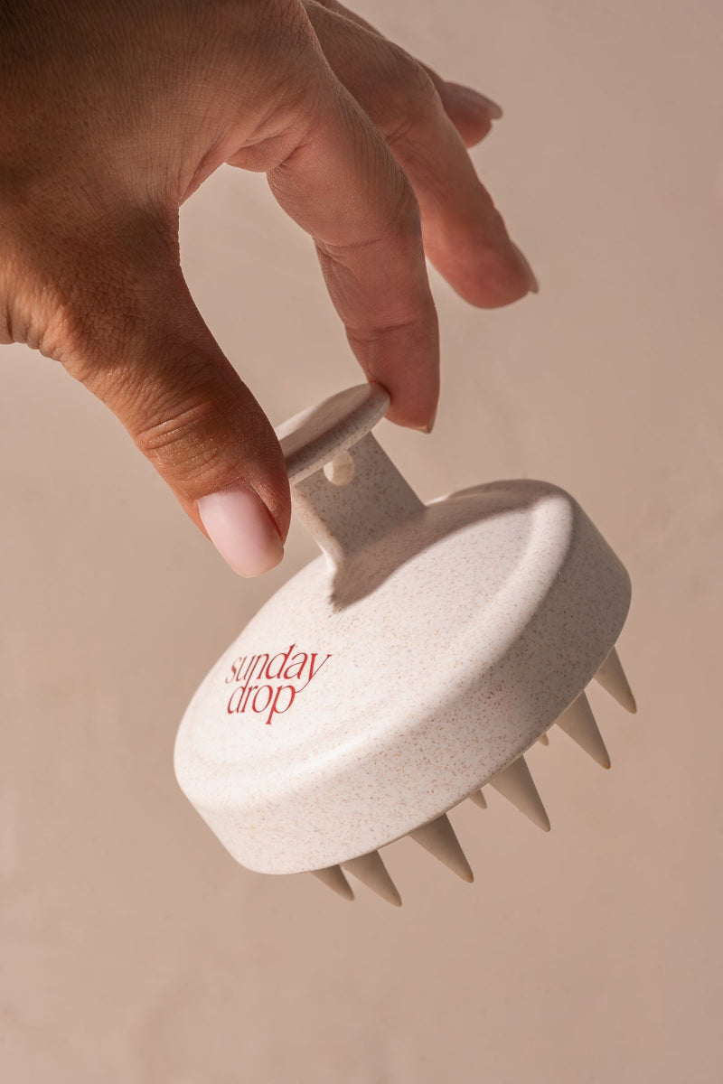 Hand holding a white scalp massager with 'sunday drop' branding on a beige background