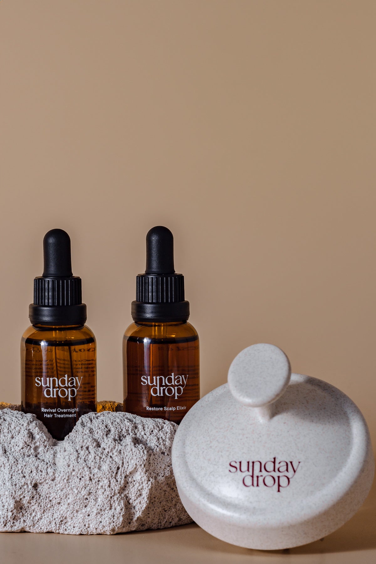 Two bottles of 'Sunday Drop' with a stone-like container on a beige background